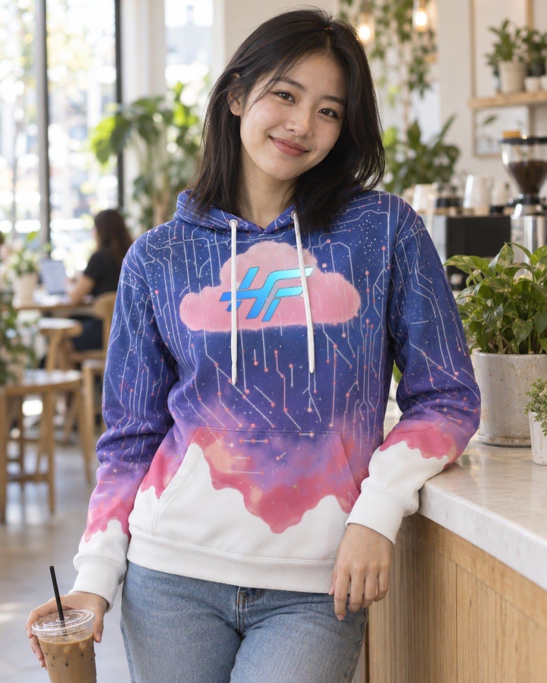 A woman with an iced coffee at a café counter, wearing the Cloud Hoodie